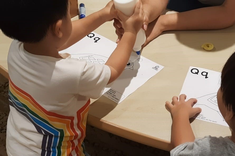 A child at School of Concepts holds a glue bottle steady with help from an adult's hand, working on a phonics craft at a table beside another child.
