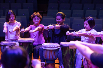 Four participants seated in a row, each drumming on a djembe during a group wellness drumming session.